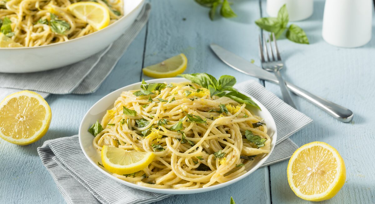 Ein Teller Zitronen-Knoblauch-Pasta mit frischen Kräutern und Zitronenscheiben, daneben Besteck, Zitronenhälften und ein weiterer Teller.