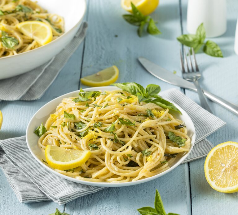 Ein Teller Zitronen-Knoblauch-Pasta mit frischen Kräutern und Zitronenscheiben, daneben Besteck, Zitronenhälften und ein weiterer Teller. | © Shutterstock/ Brent Hofacker
