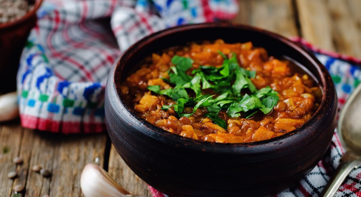 Eine Schüssel veganer Linseneintopf mit Petersilie bestreut, daneben Knoblauchzehen und Pfefferkörner.