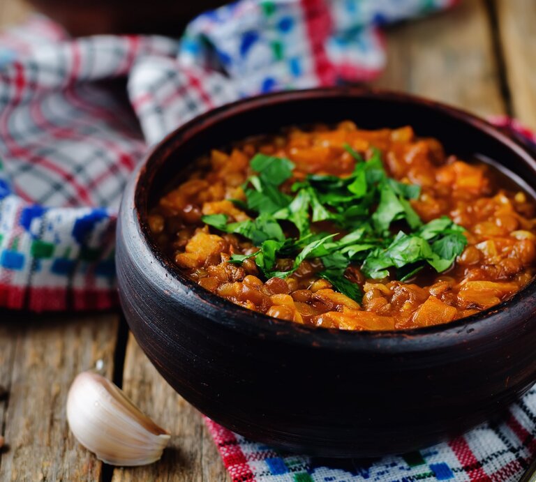 Eine Schüssel veganer Linseneintopf mit Petersilie bestreut, daneben Knoblauchzehen und Pfefferkörner. | © Adobe Stock/ nata_vkusidey