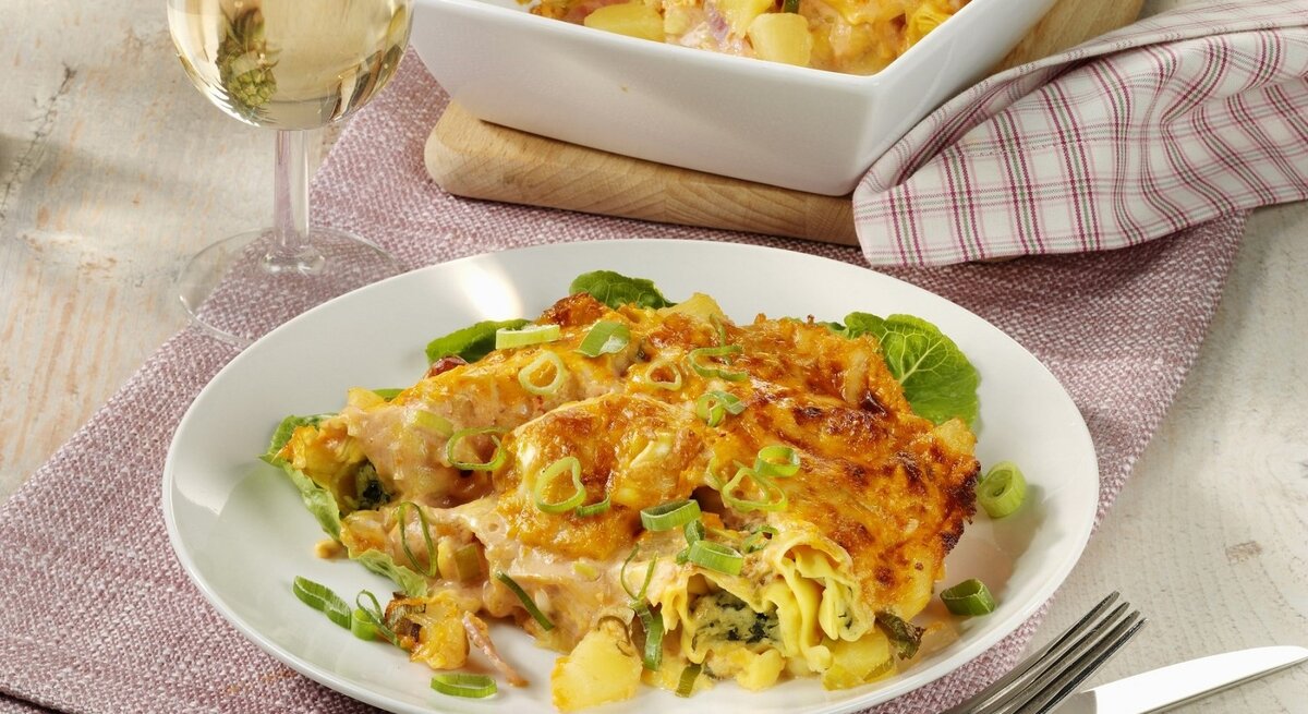 Auf einem Teller liegt eine Portion Tortellini-Auflauf 'Hawaii' mit Ananas, Schinken und Käse. Daneben steht ein Glas Weißwein.