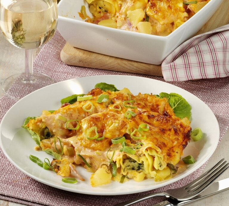 Auf einem Teller liegt eine Portion Tortellini-Auflauf 'Hawaii' mit Ananas, Schinken und Käse. Daneben steht ein Glas Weißwein. | © StockFood / StockFood Studios / Photoart