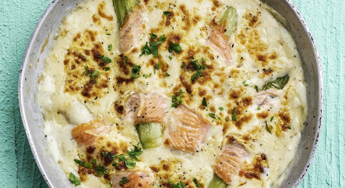 In einer Auflaufform ist ein gratinierter Lachs mit Lauch, Käsesauce und Kartoffelpüree angerichtet. Er ist mit Käse überbacken. 