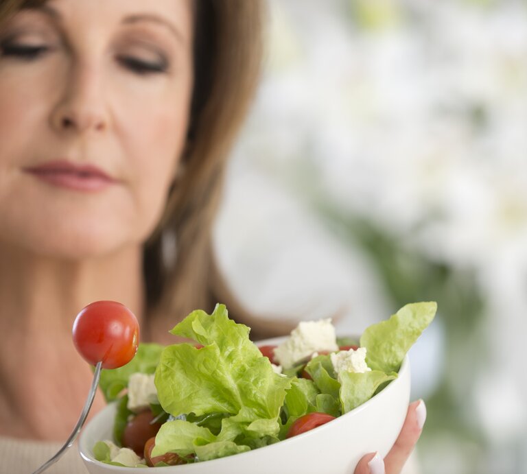 Frau mit einer Salatschale in der Hand beäugt kritisch eine kleine Tomate.  | © iStock/courtneyk