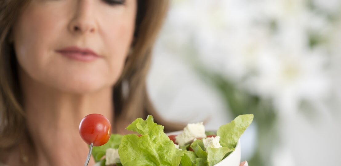 Frau mit einer Salatschale in der Hand beäugt kritisch eine kleine Tomate.  | © iStock/courtneyk