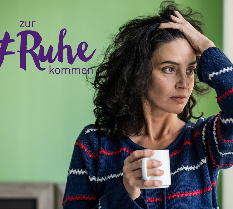 Eine nachdenkliche Frau mit dunklem Haar hält eine Tasse in ihrer Hand und blickt in die Ferne, während sie ihre andere Hand an ihren Kopf legt. Sie trägt einen gestreiften Pullover und steht in einem Raum mit grünen Wänden. Neben ihrem Kopf steht der Text "zur #Ruhe kommen" in lila Schrift, was auf einen thematischen Fokus auf Entspannung oder Selbstfürsorge hinweist. | © iStock.com/valentinrussanov