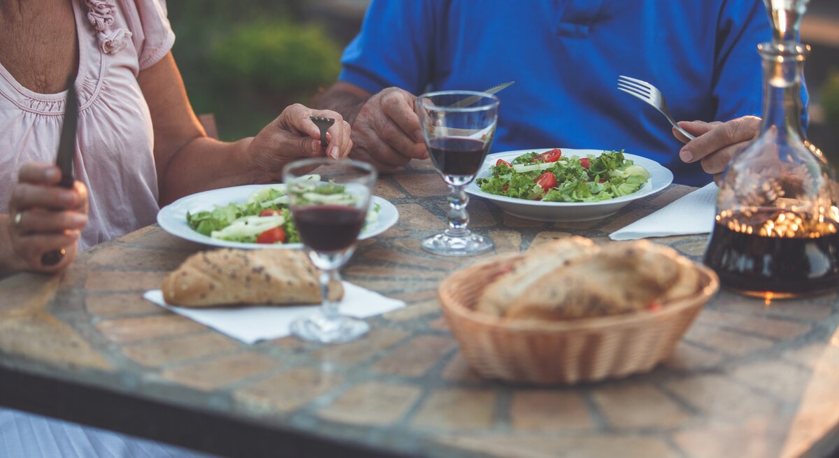 Zwei ältere Personen essen Salat und Brot an einem rustikalen Tisch im Freien, mit Rotweingläsern und einer Karaffe im Abendlicht.