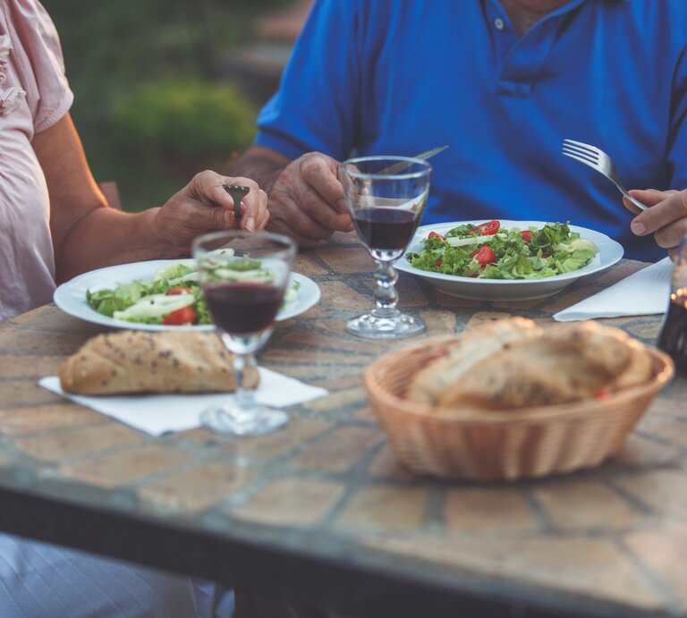 Zwei ältere Personen essen Salat und Brot an einem rustikalen Tisch im Freien, mit Rotweingläsern und einer Karaffe im Abendlicht. | © iStock.com/fotostorm
