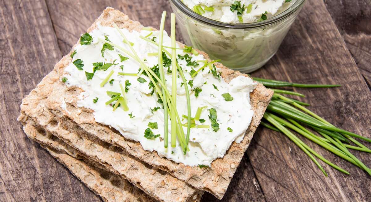 Draufsicht. Vier Scheiben Knäckebrot liegen auf einem Untergrund aus Holz. Auf der obersten Scheibe ist ein heller Aufstrich aus Frischkäse mit frischen Kräutern, wie etwa Schnittlauch und Petersilie.