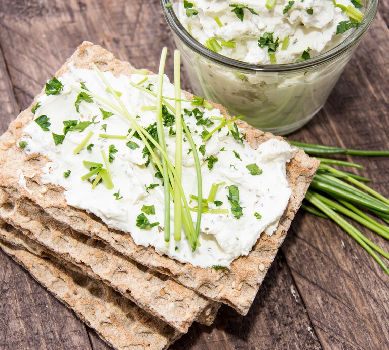 Draufsicht. Vier Scheiben Knäckebrot liegen auf einem Untergrund aus Holz. Auf der obersten Scheibe ist ein heller Aufstrich aus Frischkäse mit frischen Kräutern, wie etwa Schnittlauch und Petersilie. | © Shutterstock / HandmadePictures