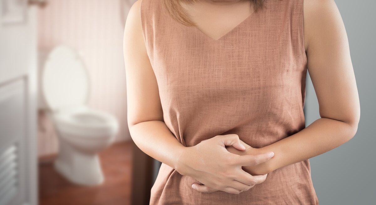 Frau hat eine Blasenentzündung und hält sich den Bauch. Im Hintergrund sieht man das Badezimmer und die Toilette. 