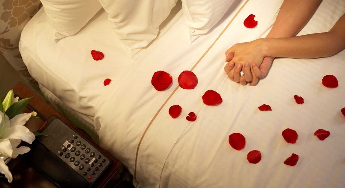 ein weißes Bett mit Rosenblüten bestreut, die verschränkten Hände eines Paares sind zu sehen