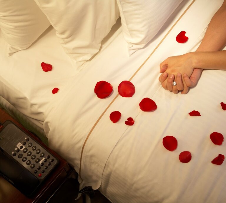 ein weißes Bett mit Rosenblüten bestreut, die verschränkten Hände eines Paares sind zu sehen | © Getty Images / blue jean images