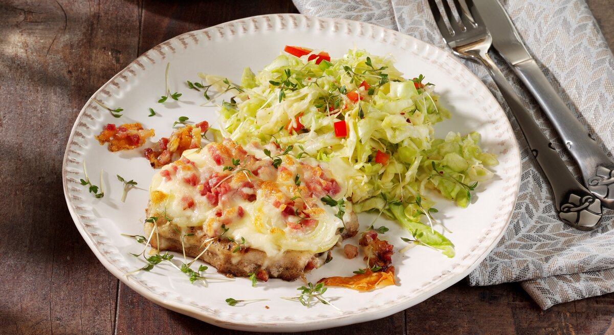 Ein weißer Teller auf einem dunklem Holztisch mit einer grau-braunen Tischdecke. Auf dem Teller ist etwas Krautsalat und das gratinierte Stück Fleisch mit Speck und Käse.
