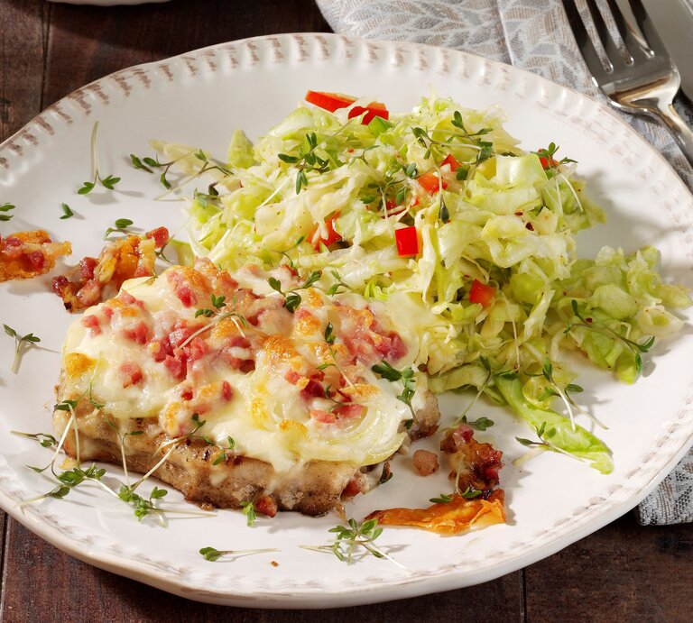 Ein weißer Teller auf einem dunklem Holztisch mit einer grau-braunen Tischdecke. Auf dem Teller ist etwas Krautsalat und das gratinierte Stück Fleisch mit Speck und Käse. | © StockFood / StockFood Studios / Photoart