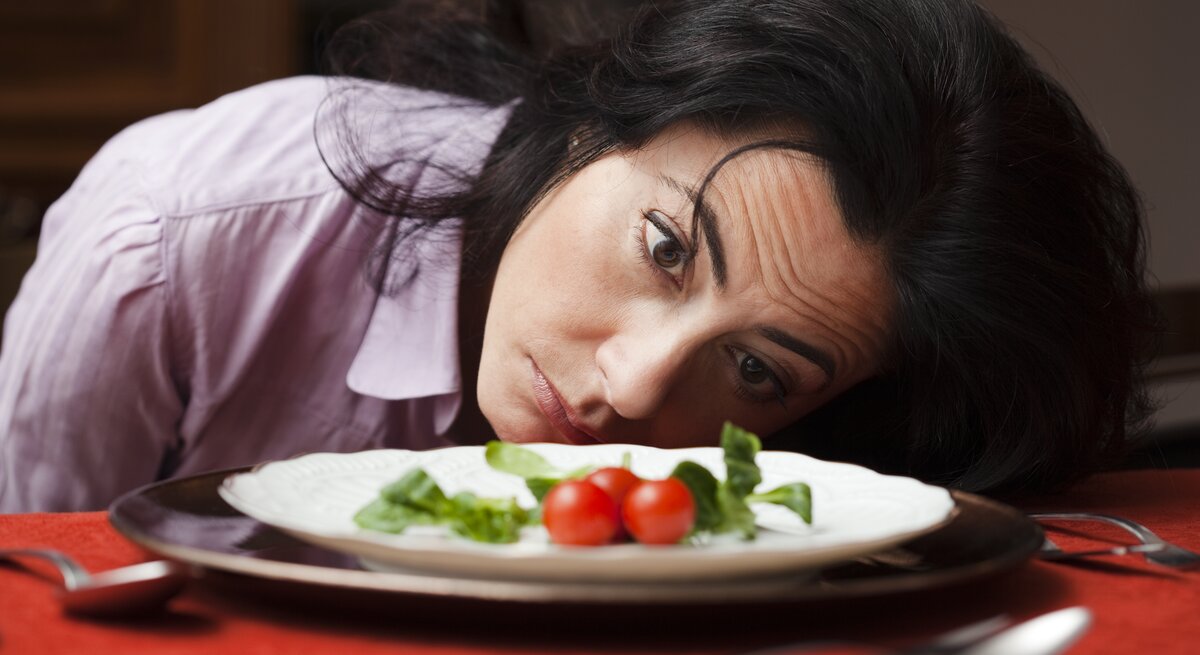 Eine Frau mittleren Alters mit dunklen Haaren und einem nachdenklichen Blick liegt mit ihrem Kopf auf einem Tisch und schaut auf einen Teller mit einer sehr kleinen Menge Essen, bestehend aus einigen Blättern Salat und zwei Kirschtomaten. Ihr Gesichtsausdruck könnte Enttäuschung, Unzufriedenheit oder Desinteresse an der spärlichen Mahlzeit ausdrücken. Sie trägt eine hellviolette Bluse, und das Ambiente wirkt häuslich. Der rote Tischläufer unter dem Teller verleiht dem Bild eine warme Atmosphäre.