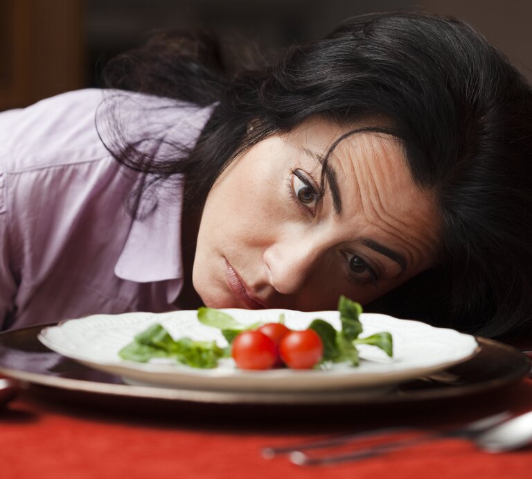 Eine Frau mittleren Alters mit dunklen Haaren und einem nachdenklichen Blick liegt mit ihrem Kopf auf einem Tisch und schaut auf einen Teller mit einer sehr kleinen Menge Essen, bestehend aus einigen Blättern Salat und zwei Kirschtomaten. Ihr Gesichtsausdruck könnte Enttäuschung, Unzufriedenheit oder Desinteresse an der spärlichen Mahlzeit ausdrücken. Sie trägt eine hellviolette Bluse, und das Ambiente wirkt häuslich. Der rote Tischläufer unter dem Teller verleiht dem Bild eine warme Atmosphäre. | © Getty Images/esolla