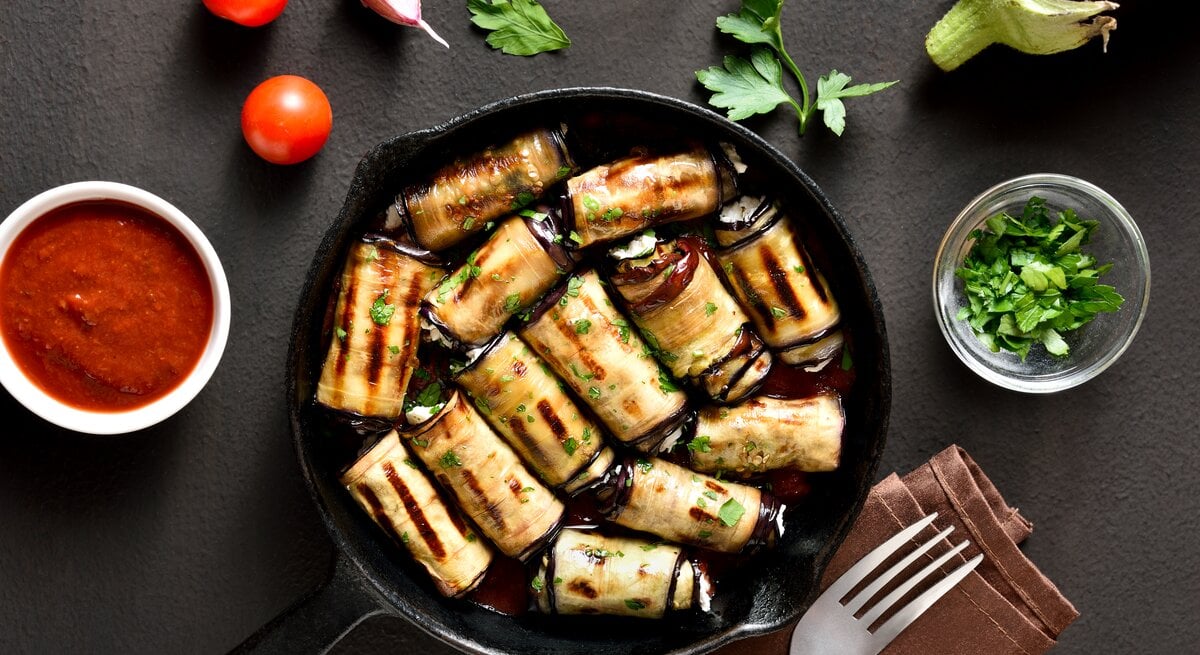 Gebratene Auberginen-Frischkäse-Röllchen in einer Pfanne. Drum herum liegen Tomaten, Petersilie, Knoblauchzehen sowie ein Dip und eine Gabel auf einem Tuch.