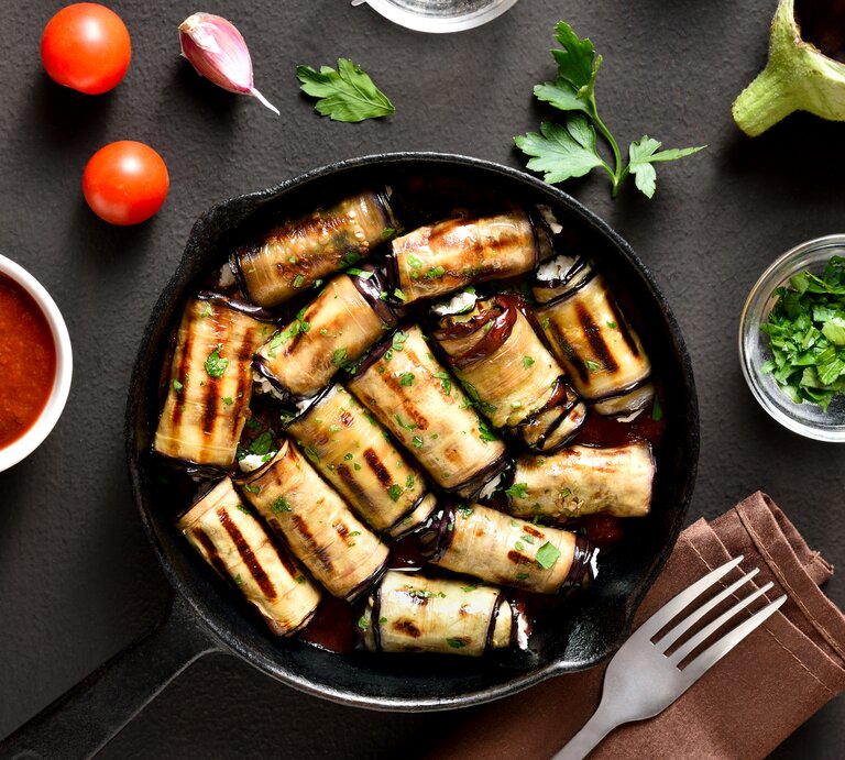 Gebratene Auberginen-Frischkäse-Röllchen in einer Pfanne. Drum herum liegen Tomaten, Petersilie, Knoblauchzehen sowie ein Dip und eine Gabel auf einem Tuch. | ©  © Shutterstock / Tatiana Volgutova