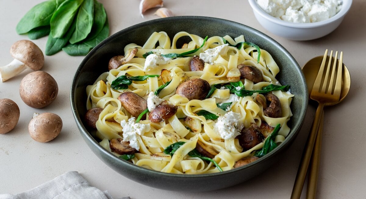 Ein grauer Teller mit dem Pastagericht auf einem hellen Tisch mit Pilzen, Spinat, Feta und Knoblauch.