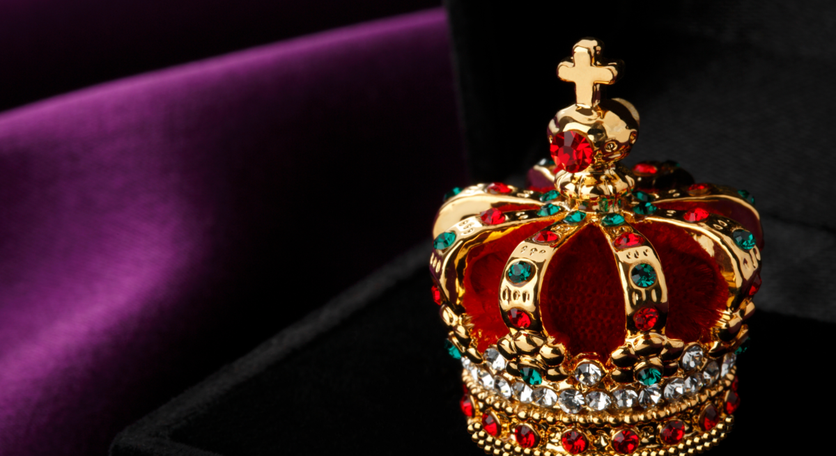 Ein prächtiges Modell einer Krone, verziert mit roten und blauen Edelsteinen sowie Diamantimitationen, ruht auf einer dunklen Samtoberfläche vor einem violett-schwarzen Hintergrund.