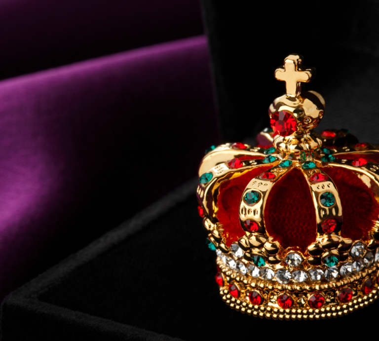 Ein prächtiges Modell einer Krone, verziert mit roten und blauen Edelsteinen sowie Diamantimitationen, ruht auf einer dunklen Samtoberfläche vor einem violett-schwarzen Hintergrund. | © Getty Images Signature/ studiocasper via Canva.com