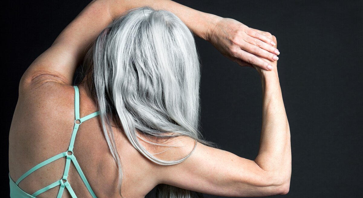Frau von hinten mit langen, grauen Haaren, die glatt herunterfallen. Man sieht auch die Schultern und einen Teil des Rückens mit türkisfarbenem Top mit gekreuztem Trägerdesign.