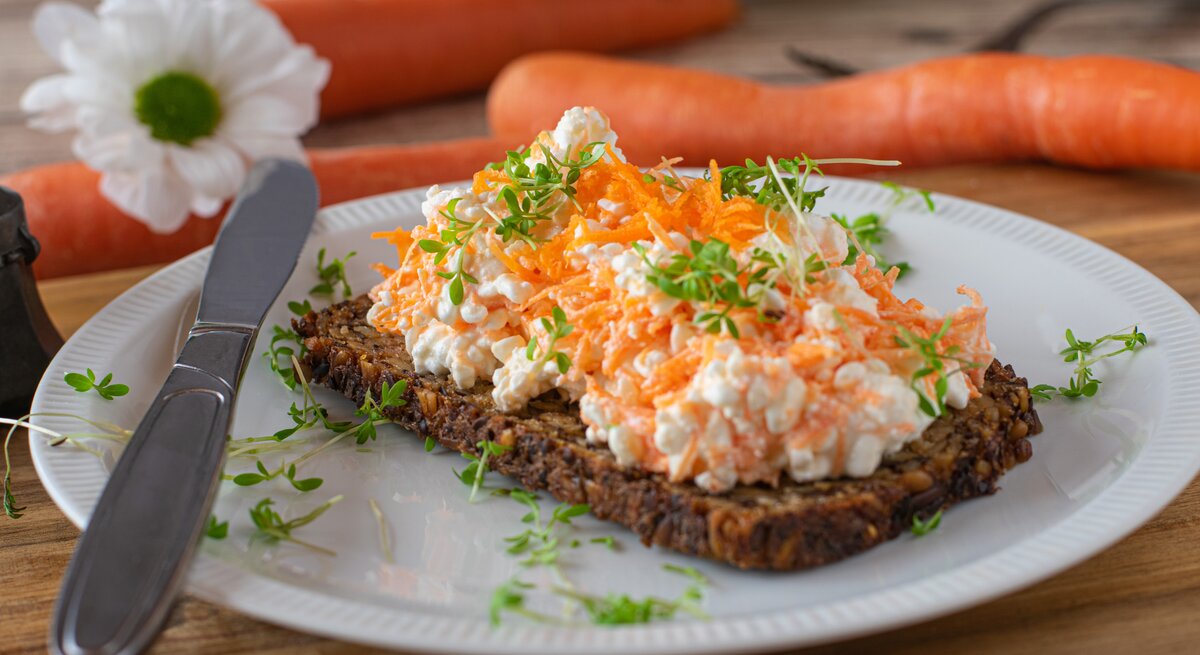 Frontal: weißer Teller mit Vollkornbrot mit Möhren-Frischkäse steht auf einem Untergrund aus Holz. Dahinter liegen drei Möhren und eine Blüte.
