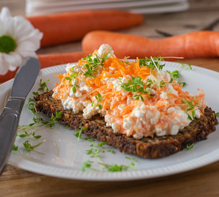 Frontal: weißer Teller mit Vollkornbrot mit Möhren-Frischkäse steht auf einem Untergrund aus Holz. Dahinter liegen drei Möhren und eine Blüte. | © Shutterstock / Angelika Heine