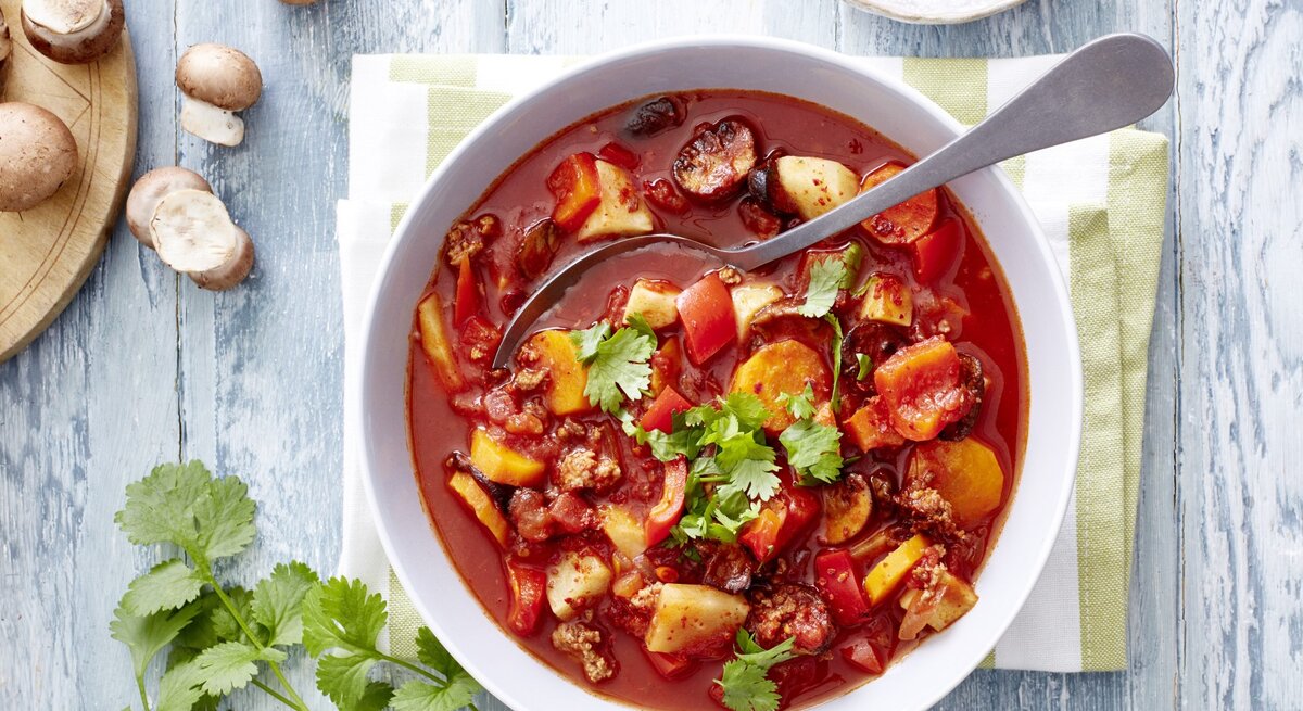 Gemüseeintopf mit Hack und Chili, bestehend aus Tomatenbrühe, Knollengemüse, kross gebratenem Hackfleisch und einer Garnitur aus Petersilie und Kroiander. Alles serviert in einer großen Suppenschale und garniert mit Chiliflocken. 