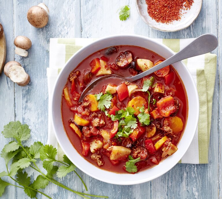 Gemüseeintopf mit Hack und Chili, bestehend aus Tomatenbrühe, Knollengemüse, kross gebratenem Hackfleisch und einer Garnitur aus Petersilie und Kroiander. Alles serviert in einer großen Suppenschale und garniert mit Chiliflocken.  | © EAT CLUB / Isabell Triemer