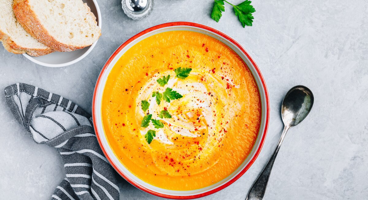 Ein Teller mit Zucchini-Möhren-Suppe in der Draufsicht. Daneben steht eine Schale mit Baguettescheiben, Gewürzstreuer und ein Löffel.