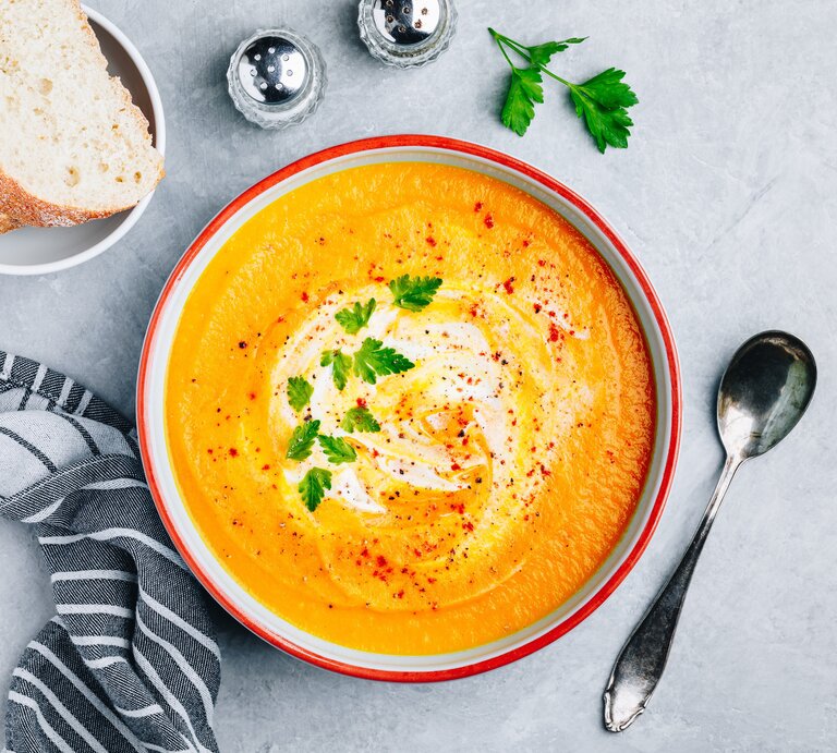 Ein Teller mit Zucchini-Möhren-Suppe in der Draufsicht. Daneben steht eine Schale mit Baguettescheiben, Gewürzstreuer und ein Löffel. | © Getty Images/ wmaster890