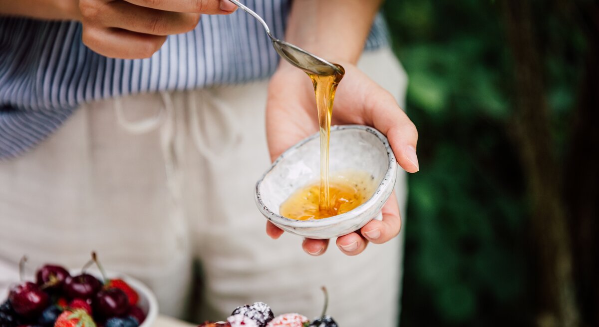 Eine Person gießt mit einem Löffel Reissirup aus einer kleinen Schale. Im Hintergrund unscharf eine Schüssel mit verschiedenen frischen Beeren.