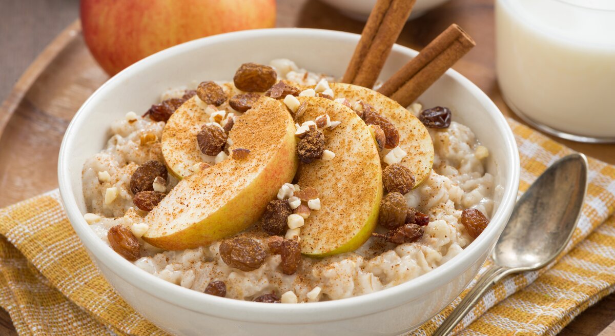 Eine weiße Schüssel mit Apfelstrudel-Porridge neben einem Löffel auf einem gelb-weiß-karierten Geschirrtuch.