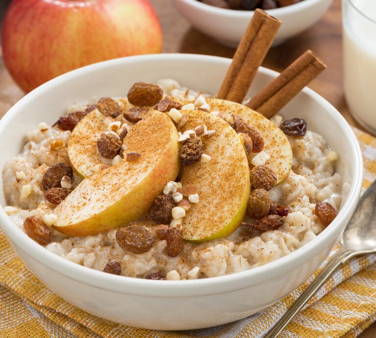 Eine weiße Schüssel mit Apfelstrudel-Porridge neben einem Löffel auf einem gelb-weiß-karierten Geschirrtuch. | © Shutterstock / Yulia Davidovich