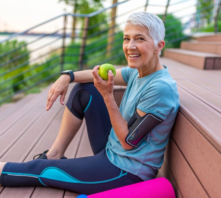 Ältere Frau sitzt nach dem Joggen auf einer Tribüne und isst einen Apfel.  | © Shutterstock/Dragana Gordic