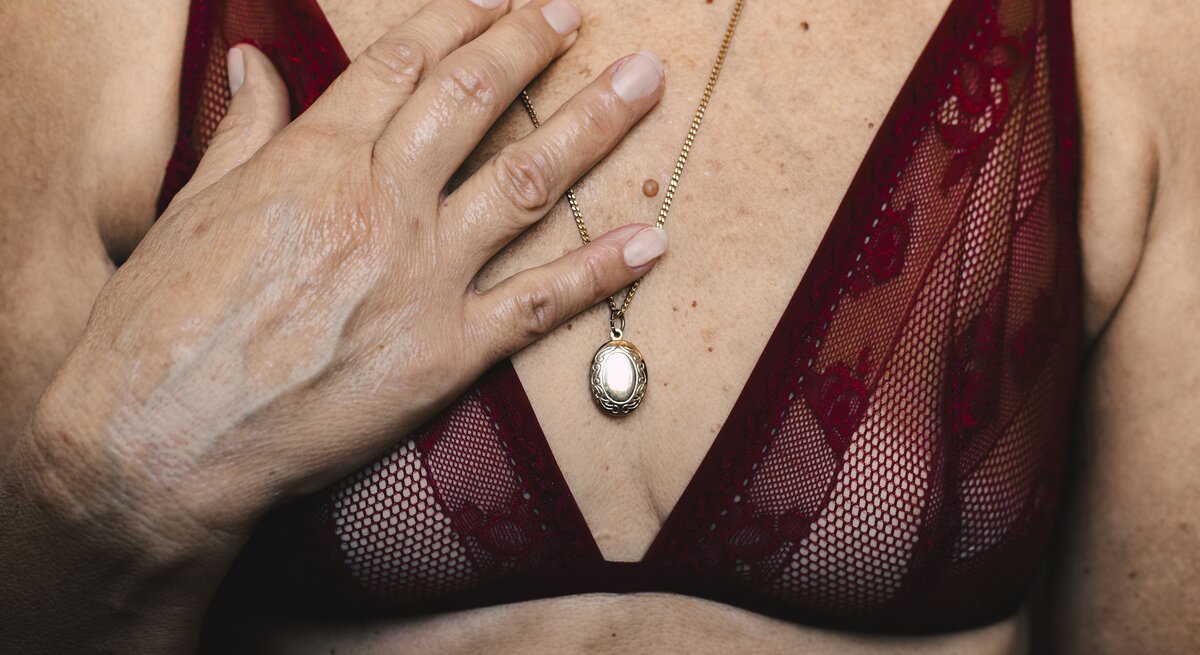 Das Bild zeigt einen nahen Ausschnitt einer Frau in roter Spitzenunterwäsche. Sie hat eine Hand auf ihrer Brust und trägt eine goldene Kette mit einem ovalem Anhänger. Der Fokus liegt auf der goldenen Kette gegen den roten Stoff, was einen starken Kontrast bildet. Die Haut ist natürlich gealtert, was die natürliche Schönheit und Eleganz im Alter unterstreicht.