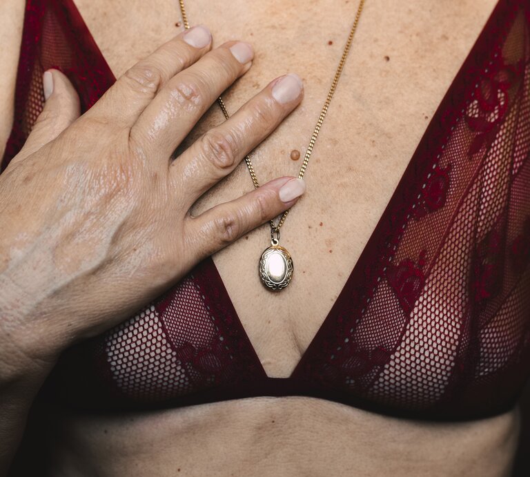 Das Bild zeigt einen nahen Ausschnitt einer Frau in roter Spitzenunterwäsche. Sie hat eine Hand auf ihrer Brust und trägt eine goldene Kette mit einem ovalem Anhänger. Der Fokus liegt auf der goldenen Kette gegen den roten Stoff, was einen starken Kontrast bildet. Die Haut ist natürlich gealtert, was die natürliche Schönheit und Eleganz im Alter unterstreicht. | © Getty Images /	Westend61