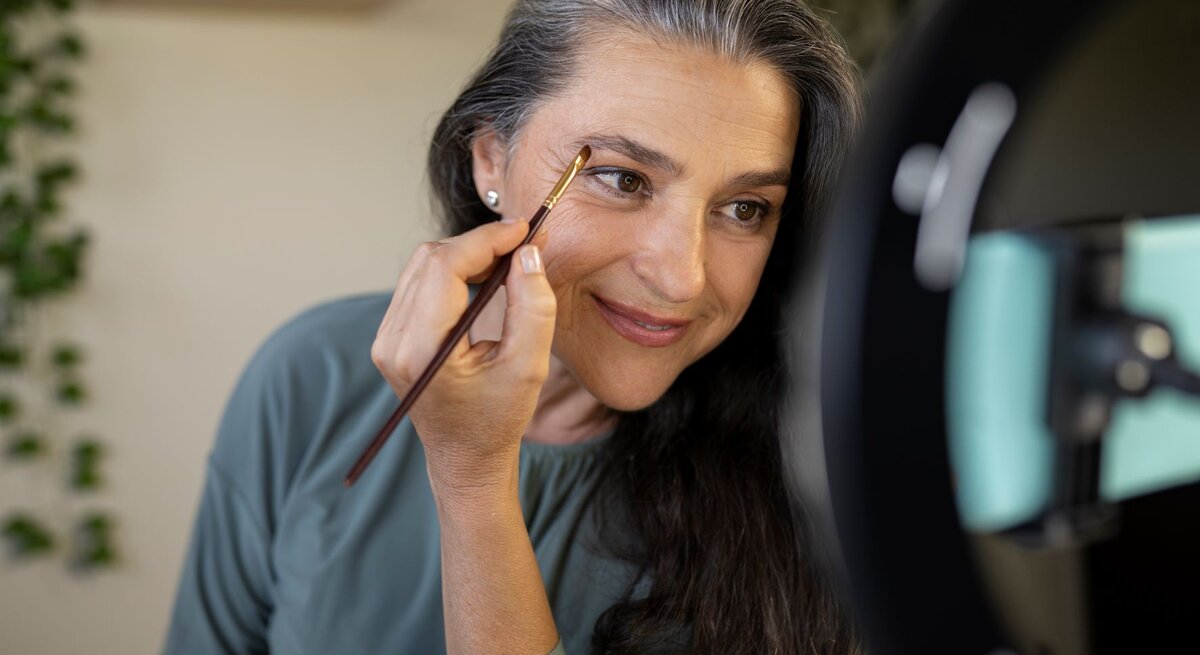 Das Foto zeigt eine Frau mittleren Alters mit graumelierten Haaren, die sich mit einem Make-up-Pinsel die Augenbrauen nachzieht. Sie blickt dabei in einen beleuchteten Kosmetikspiegel. Ihr freundliches Lächeln und der Blickkontakt mit ihrem Spiegelbild vermitteln eine positive und entspannte Stimmung. Sie trägt ein schlichtes, türkisfarbenes Top, das ihre natürliche Schönheit unterstreicht. Die Szenerie deutet auf eine alltägliche Schönheitsroutine hin.