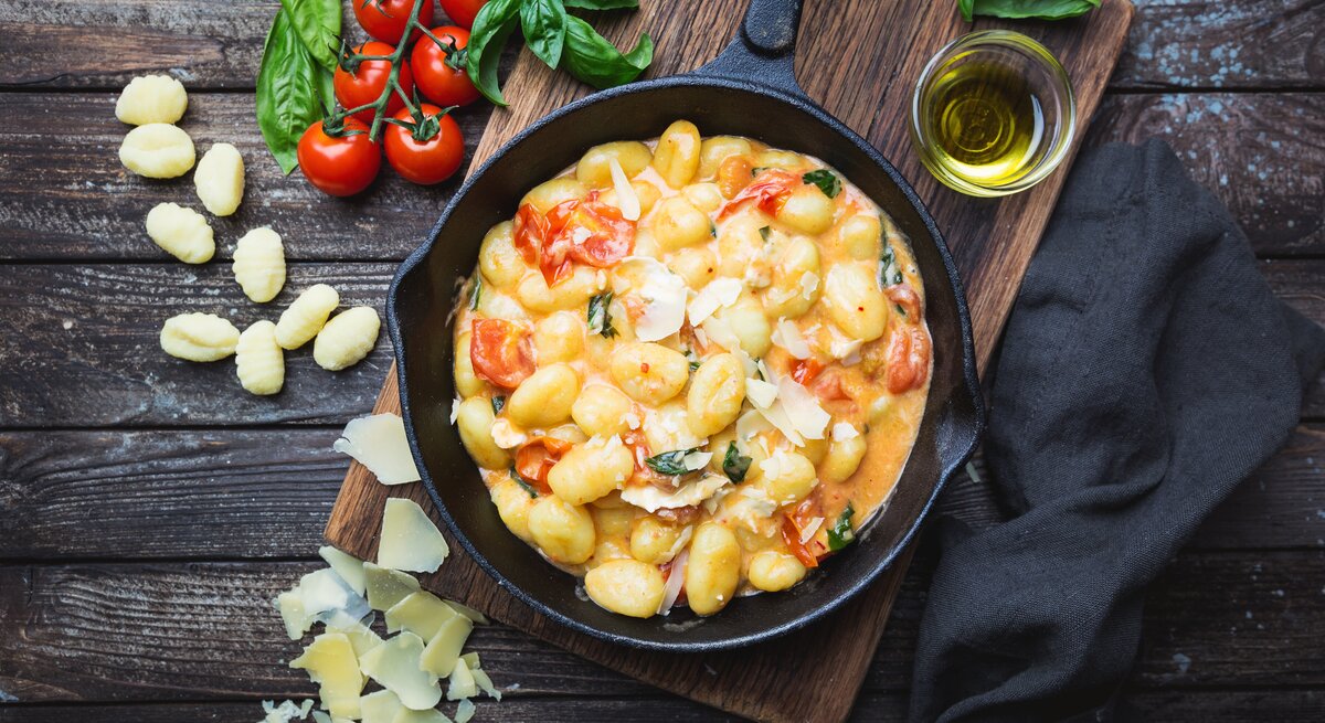Gnocchi in Tomaten-Rahm-Soße in gusseiserner Pfanne auf Holztisch. Daneben liegen Tomaten und ungekochte Gnocchi.