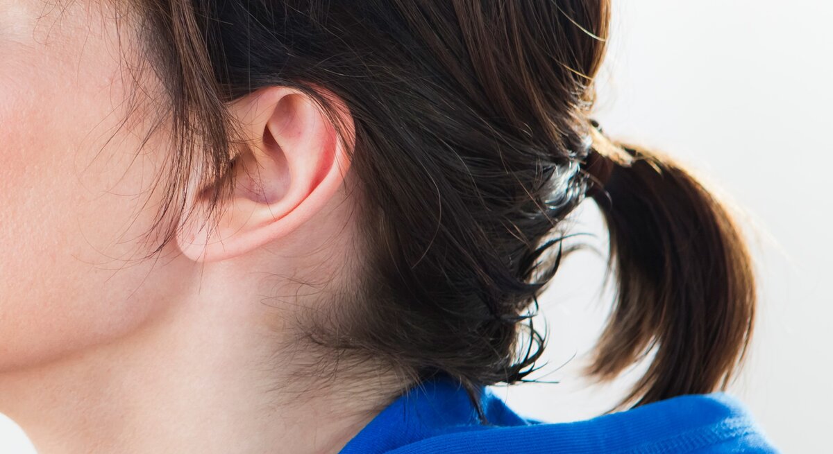 Teils eines weiblichen Kopfes mit Fokus auf dem Ohr und den Haaren. Das Ohr ist vollständig sichtbar und die Haare sind zu einem kleinen Pferdeschwanz gebunden, der mit einem unauffälligen Haargummi zusammengehalten wird.