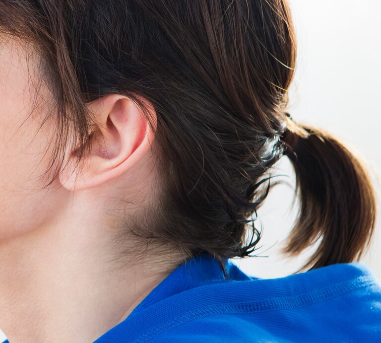 Teils eines weiblichen Kopfes mit Fokus auf dem Ohr und den Haaren. Das Ohr ist vollständig sichtbar und die Haare sind zu einem kleinen Pferdeschwanz gebunden, der mit einem unauffälligen Haargummi zusammengehalten wird. | © Getty Images / Sporrer/Rupp