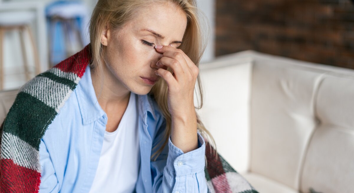 Zu sehen ist eine blonde Frau, die auf einem Sofa sitzt. Sie trägt ein hellblaues Hemd und darunter ein weißes Shirt. Sie hat eine Decke um ihre Schultern gelegt, und reibt mit der Hand zwischen ihren Augen, über der Nase. Die Augen sind geschlossen, die Frau scheint müde und erschöpft. 