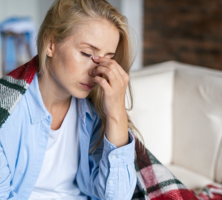 Zu sehen ist eine blonde Frau, die auf einem Sofa sitzt. Sie trägt ein hellblaues Hemd und darunter ein weißes Shirt. Sie hat eine Decke um ihre Schultern gelegt, und reibt mit der Hand zwischen ihren Augen, über der Nase. Die Augen sind geschlossen, die Frau scheint müde und erschöpft.  | © Getty Images/ brizmaker