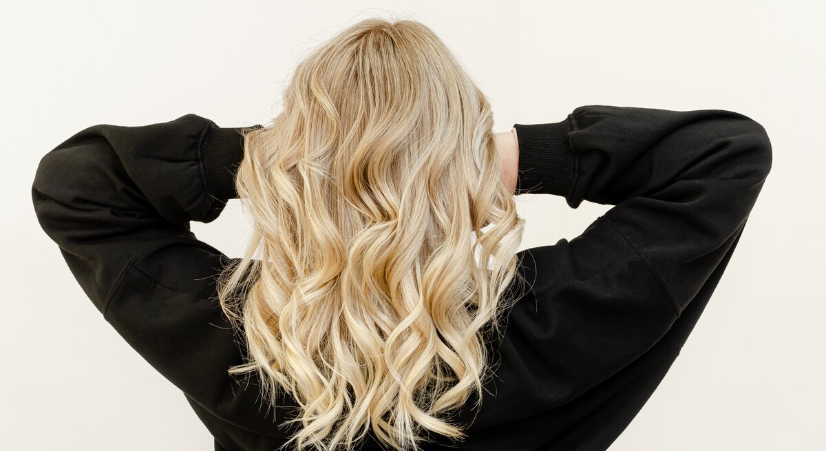 Eine Frau mit blonden Haaren steht mit dem Rücken zur Kamera und fasst in ihre Haare. Sie trägt ein schwarzes Oberteil.