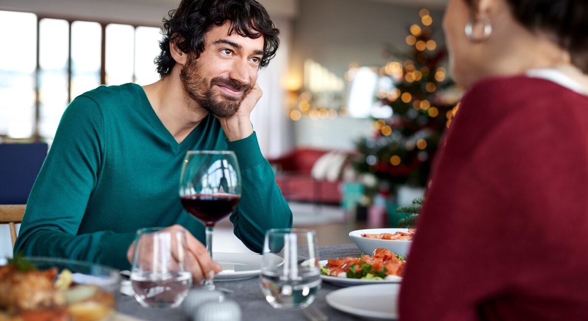 Ein dunkelhaariger Mann in einem grünen Pullover sitzt an einem reichlich gedeckten Tisch und schaut eine dunkelhaarige Frau, die ihm gegenüber sitzt, mit einem liebevollen und zufriedenen Lächeln an. Vor ihm steht ein Glas Rotwein. Der Hintergrund ist unscharf gehalten.