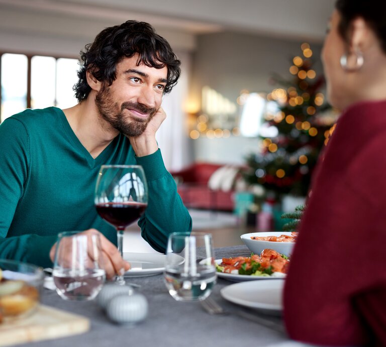 Ein dunkelhaariger Mann in einem grünen Pullover sitzt an einem reichlich gedeckten Tisch und schaut eine dunkelhaarige Frau, die ihm gegenüber sitzt, mit einem liebevollen und zufriedenen Lächeln an. Vor ihm steht ein Glas Rotwein. Der Hintergrund ist unscharf gehalten. | © Getty Images / 	Morsa Images