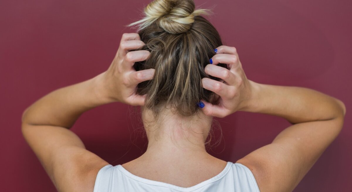 Rückansicht einer Frau, die sich mit beiden Händen an den Kopf fasst. Sie trägt ein weißes Tanktop und hat ihr blondes Haar zu einem lässigen Dutt hochgesteckt. Ihre Fingernägel sind blau lackiert. Der Hintergrund ist einheitlich weinrot, was einen Kontrast zu ihrer hellen Kleidung und Haarfarbe bildet.