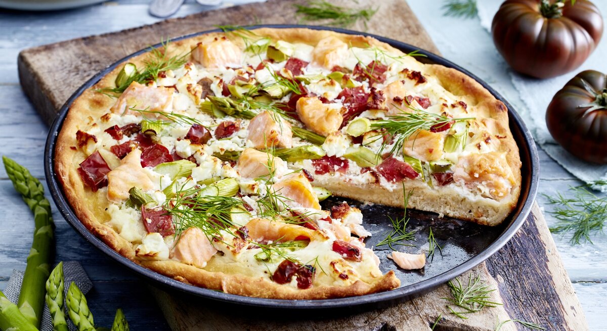 Angeschnittene Lachs-Feta-Pizza auf dunklem Teller. Drum herum Gemüse auf hellem Holzuntergrund. 